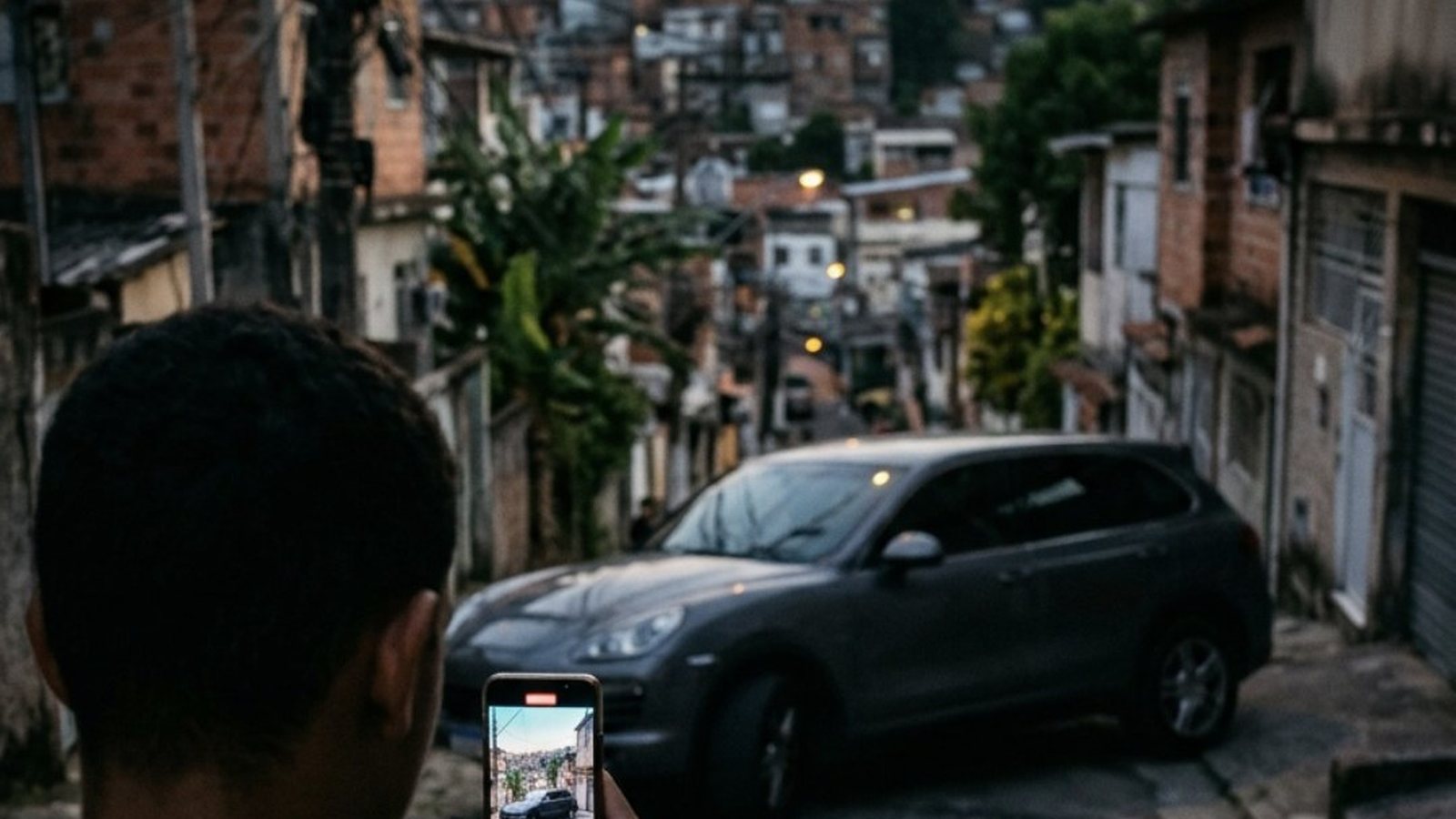 SUV de luxo cinza escuro estacionado numa rua estreita de morro no Rio de Janeiro ao entardecer, filmado por um jovem que segura o celular na vertical, visto por trás do ombro, com casas e fios desfocados ao fundo