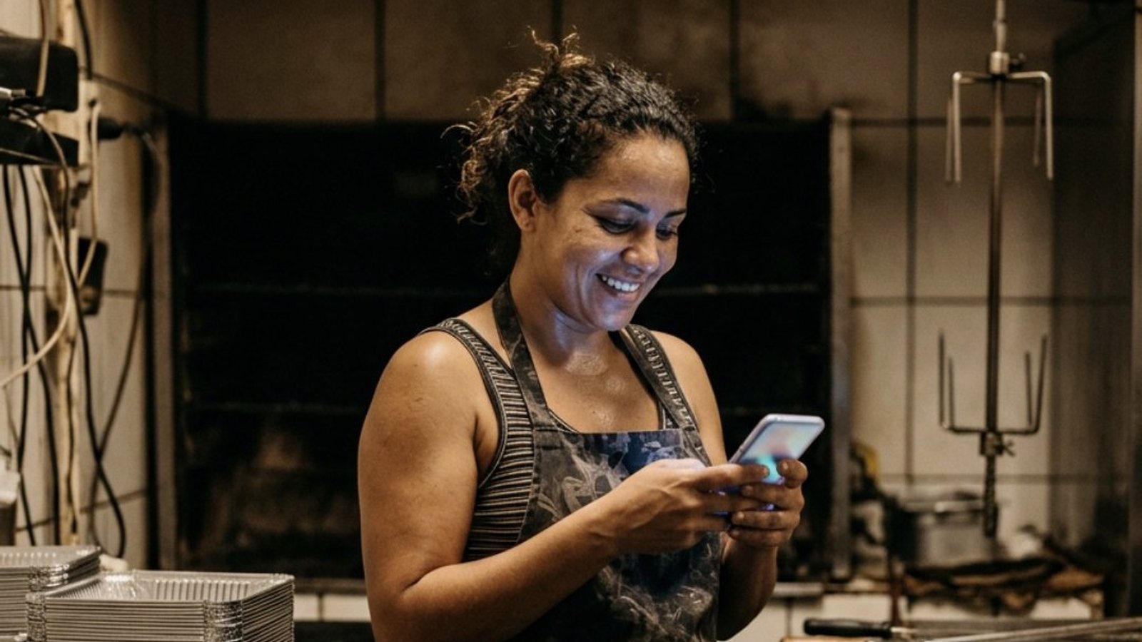 Mulher de quase quarenta anos na cozinha de uma pequena casa de frango assado, de pé diante do espaço vazio onde ficaria um forno a gás, olhando o celular com um sorriso de expectativa, entre bancadas de aço e bandejas de alumínio