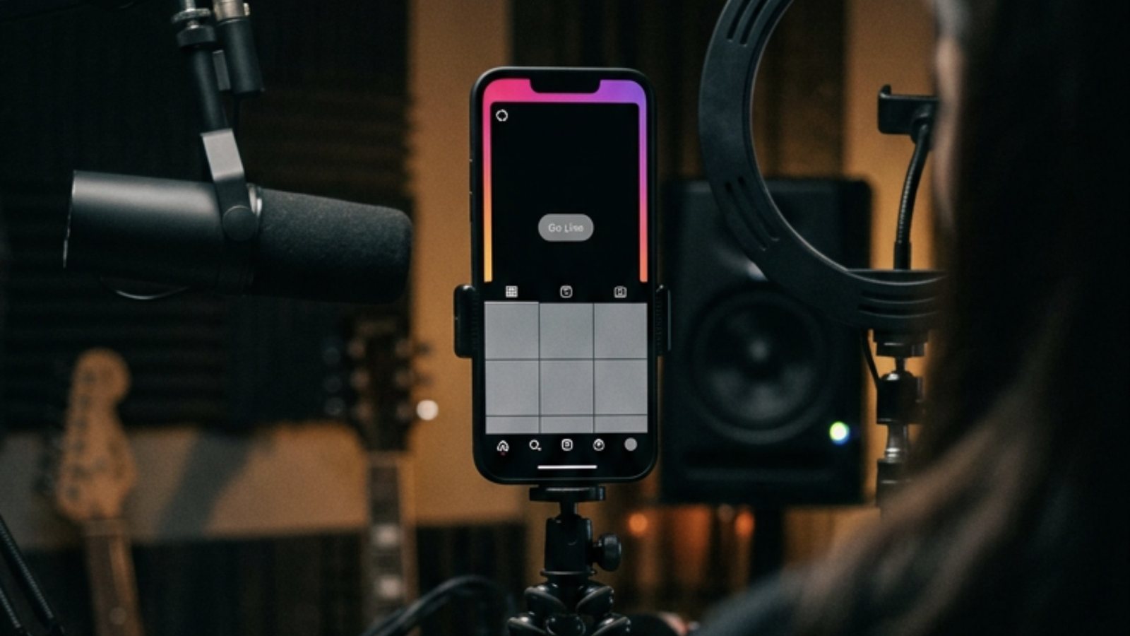 Celular preso num tripé em um estúdio escuro, com a tela de degradê rosa e roxo, o botão Go Live apagado em cinza e a grade de fotos vazia, entre um microfone e um anel de luz desligado
