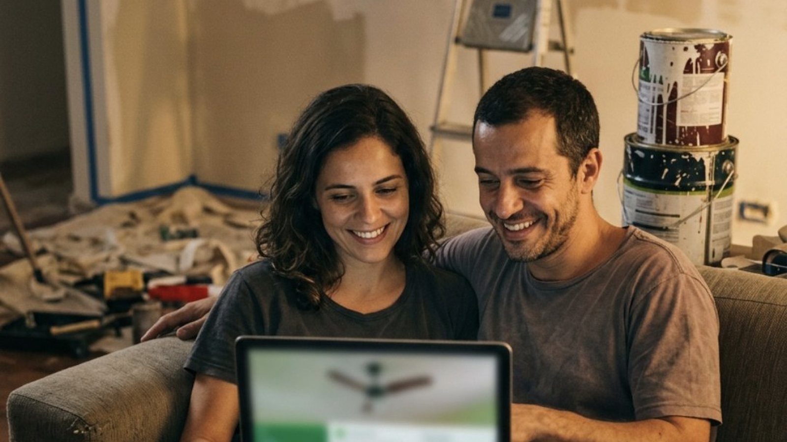 Casal brasileiro de uns trinta anos sentado no sofá de uma sala em reforma, com latas de tinta e uma escada ao fundo, a mulher segurando um notebook com uma loja virtual de detalhes verdes e a foto grande de um ventilador de teto, os dois sorrindo com o preço, sob luz de abajur
